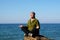 Middle aged man sits in lotus position on a large rock by the sea and meditates in sunlight, solitude in nature