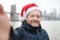 Middle aged man tourist in Santa Claus hat takes selfie in front of stunning views of Brooklyn Bridge and Manhattan skyscrapers