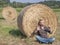 A middle-aged white man listens to music on headphones sitting in a field, with his back to a bale of hay