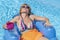 A middle-aged white woman relaxes lying on an inflatable mattress on the water of a swimming pool, with a glass of cocktail in her