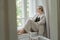 A middle-aged woman in a beige suit and black tank top sits mysteriously and looks out the window on the windowsill