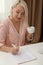 Middle aged woman with cup of drink solving sudoku at table indoors