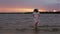 Middle aged woman in a dress and sun hat is walking barefoot in the water at sunset.