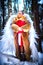 A middle-aged woman in a large warm fur coat, red Santa hat, big bag of gifts and white angel wings in a winter forest with snow