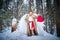 A middle-aged woman in a large warm fur coat, red Santa hat, big bag of gifts and white angel wings in a winter forest with snow