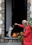 A middle-aged woman in a red cloak with a net of orange pumpkins opens the door