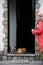 A middle-aged woman in a red cloak with a net of orange pumpkins opens the door