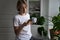 Middle-aged woman scrolls social media on smartphone holding mug with coffee near pot-plants