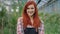 In the middle of flower greenhouse pretty redhead lady gardener posing in front of the camera smiling large showing a