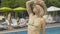 Middle shot of confident blond woman in golden suit waving sun hat at poolside. Portrait of beautiful adult Caucasian