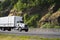 Middle size semi truck with long box trailer transporting flammable commercial cargo running on the green road with rocks