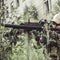 A military man or airsoft player in a camouflage suit sits in the grass and aims from an automatic rifle to the side