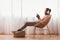 Millennial Afro Guy In Headphones Using Smartphone Sitting Indoor