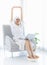 Millennial Asian sleepy female in white clean bathrobe and towel waking up sitting on cozy armchair stretching arms and hands up