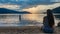 Millstaettersee - A woman in a blue dress sitting at the shore of Millstaettersee lake and enjoying the sunset. The sun is setting