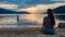 Millstaettersee - A woman in a blue dress sitting at the shore of Millstaettersee lake and enjoying the sunset. The sun is setting