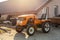 Mini red tractor with trailer standing near hangar building at farm during sunset or sunrise. Small agricultural