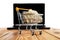 Miniature supermarket shopping cart full of shipping boxes stand on a wooden table against a blurred black laptop on white