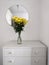 A minimalist bedroom interior, a guest room with a round mirror, white chest of drawers with yellow chrysanthemums in a vase