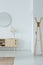 Mirror above stylish wooden cupboard with glass vase and flower on it, modern clothes hanger in the corner of white hall