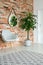 Mirror on the wall of corridor with tall plant in pot, grey stylish chair and brick wall
