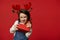 Mischievous funny little child girl in deer antler hoop, unpacking gift box with surprise for Christmas, red background