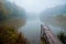 Misty autumn morning on a small lake, fog weather, wooden footbridge over still water surface, yellow trees reflections