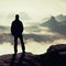 Misty day in rocky mountains. Silhouette of tourist with poles in hand. Hiker stand on rocky view point above misty valley.