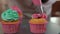 Mixed pink and green royal icing pouring from pastry bag on baked muffin indoors. Unrecognizable woman decorating