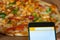 Mobile phone in a white and yellow touchscreen with a chicken and peppers pizza in a noisy background out of focus