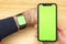 Mockup image of hands holding black mobile phone with green screen and electronic smart watch on wrist . Office table on the.