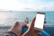 Mockup image of a man`s hand holding white mobile phone with blank desktop screen sitting by the sea and blue sky