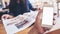 Mockup image of a man`s hand holding white mobile phone with blank screen in modern cafe and blur woman reading newspaper