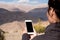 Mockup image of a woman using white mobile phone with blank black screen while standing in front of mountain