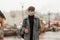 Model of a fashionable young man in an elegant youth checkered coat with a trendy hairstyle stands in the city square with a hot