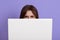 Model hiding behind laptop isolated over lilac background, having angry facial expression, dark haired female behind white note
