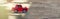 A model of a red toy car in the sunbeams on the road rides outdoors. Close-up. Horizontal format banner with place for