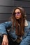 Modern American young hipster woman in stylish purple glasses in youth blue denim jacket is relaxing outdoors near gray building.