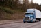 Modern blue powerful stylish big rig semi truck with semi trailer going on wide highway with trees on the hillside shoulder