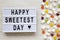 Modern board with `Happy Sweetest Day` word and candy on a white wooden background, overhead view. Top view, flat lay, from abov