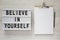 Modern board with text `Believe in yourself`, clipboard with sheet of paper on a white wooden background, top view. From above,