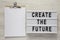 Modern board with text `Create the future`, clipboard with sheet of paper on a white wooden background, top view. From above, fl