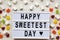 Modern board with text `Happy Sweetest Day` word and candy over white wooden background, overhead view. From above, flat lay