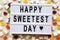 Modern board with text `Happy Sweetest Day` word and candy over white wooden surface, top view. From above, flat lay