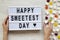 Modern board with text `Happy Sweetest Day` word and female hands with candy over white wooden background, top view. From above,