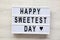 Modern board with text `Happy Sweetest Day` word over white wooden surface, top view. From above, flat lay