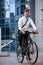 Modern building behind. Businessman in formal clothes with black bicycle is in the city