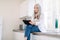 Modern charming senior lady with long gray hair, wearing jeans and white shirt, sitting on the kitchen countertop while