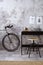 Modern composition of indiustral concrete office interior with black desk, stylish stool, bike, books, gray concrete wall, boho