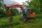 A modern excavator stands on the grass. The bucket is lowered. In the background there are trees and a village house